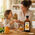 Mutter reicht ihrem Kind am Küchentisch einen Messbecher Riviera Immunkomplex mit Propolis und Honig, daneben Honigwabe und Orangen | Naturpräparate Dieterich