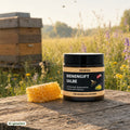 Riviera Bienengiftsalbe auf einem Holztisch neben einer Honigwabe, im Hintergrund ein Bienenstock in der Wiese | Naturpräparate Dieterich 